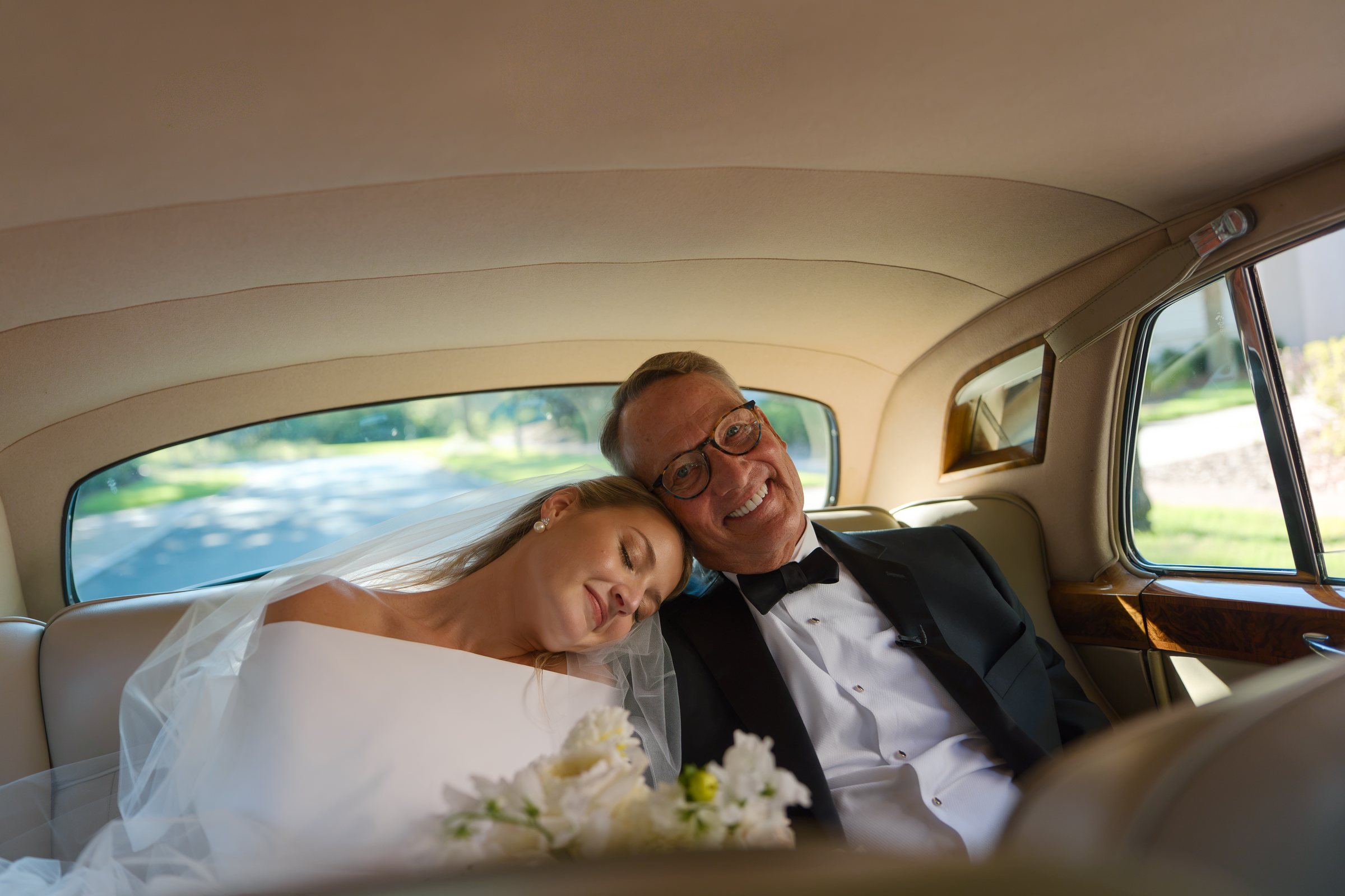 A bride resting her head on his shoulder in the back of a vintage car